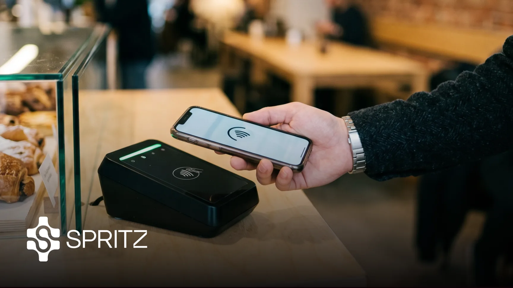 A customer taps their phone to pay at a cafe counter, with the Spritz logo